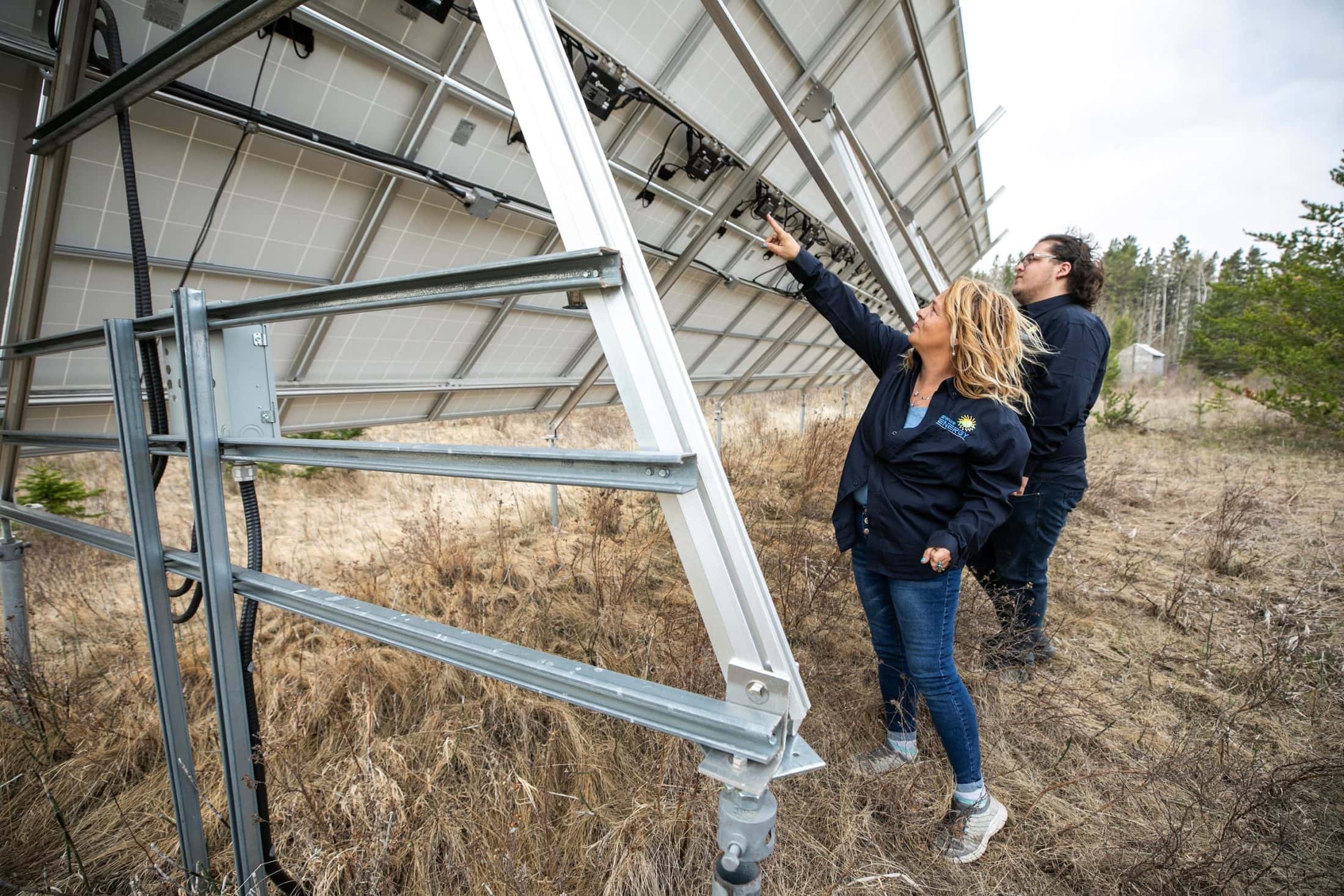 Two people stand under a large solar panel. One points at wiring on the back of the panel