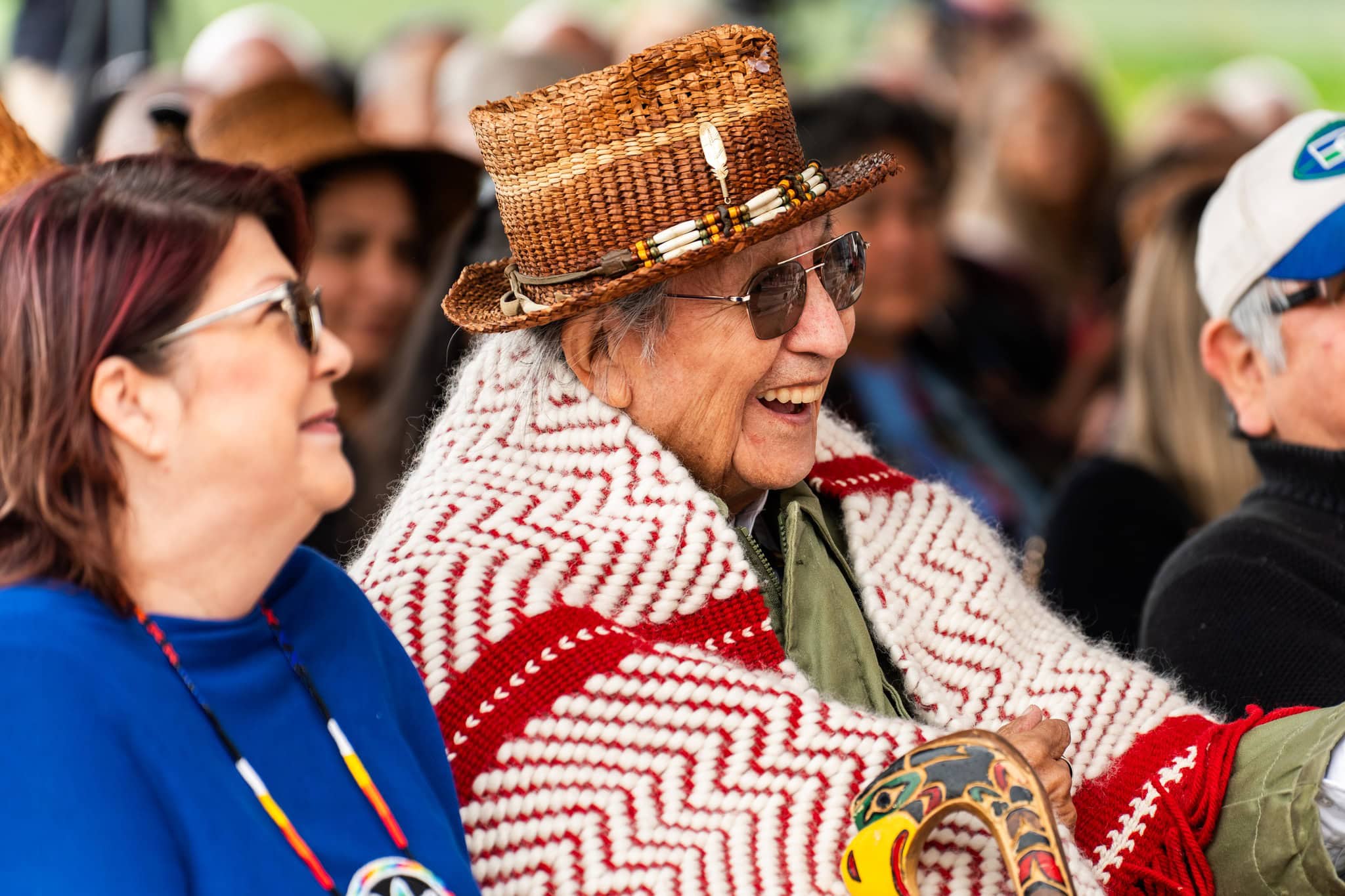 A man wearing a cedar hat smiles wrapped in a blanket.