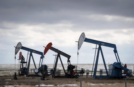 Three oil pumpjacks sit on a pad in a vast prairie field, partly covered in snow