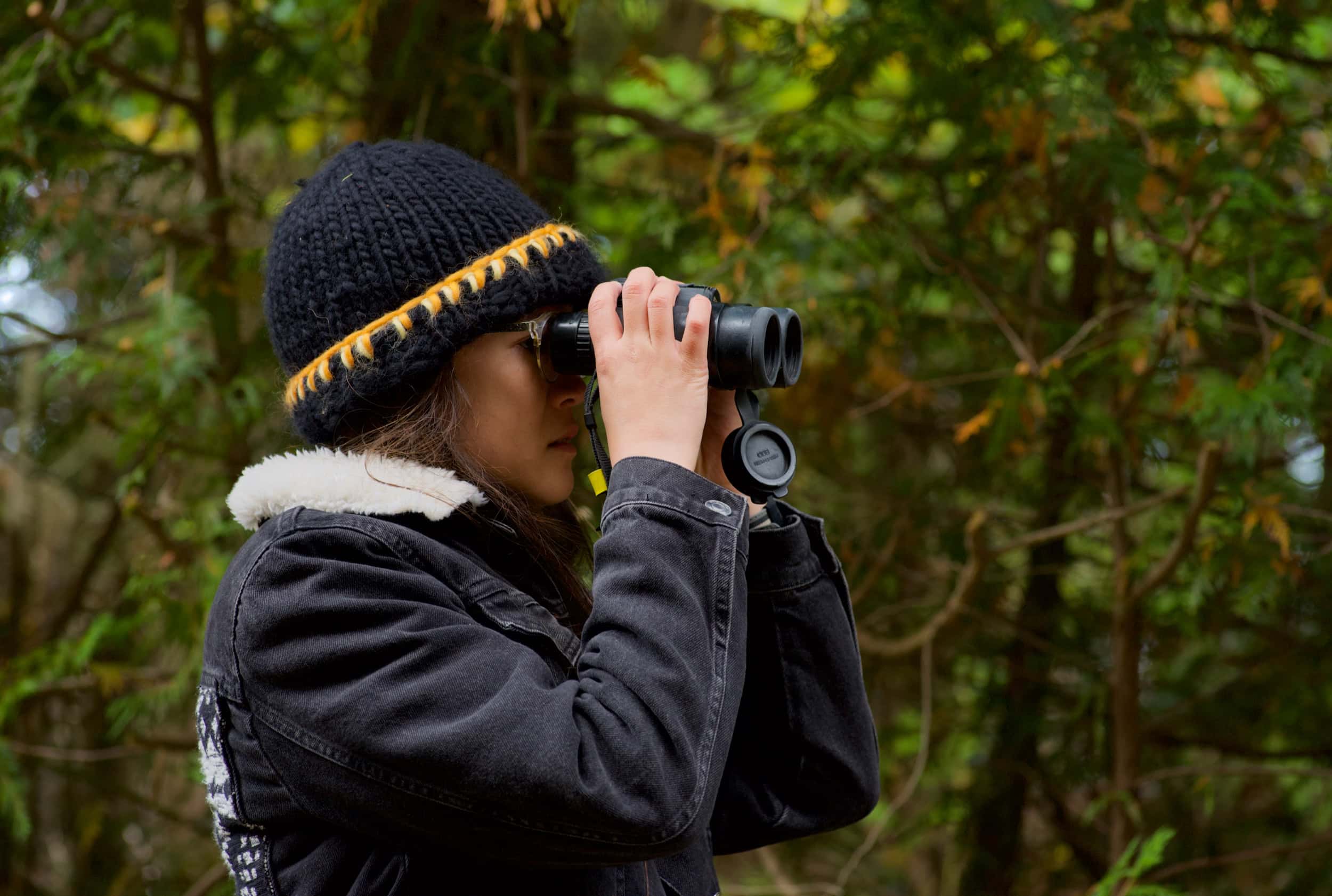 A woman looking into binoculars with a forest in the backdrop.