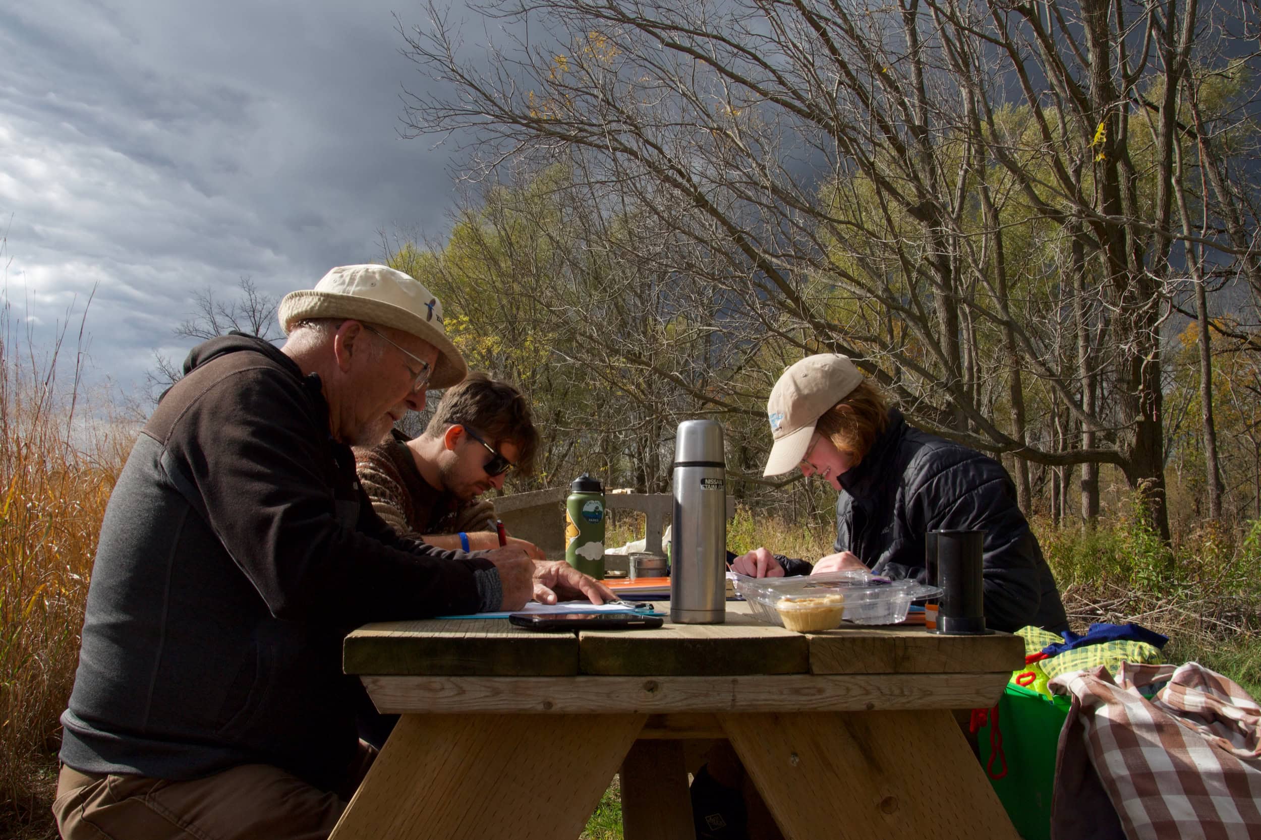 Three people sit at a wooden picnic bench, working in notebooks.