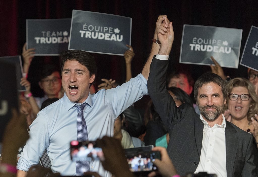 Canadian politicians Justin Trudeau and Steven Guilbeault raise their hands together during a campaign event in 2019. Supporters hold signs that read, "ÉQUIPE TRUDEAU" behind them.