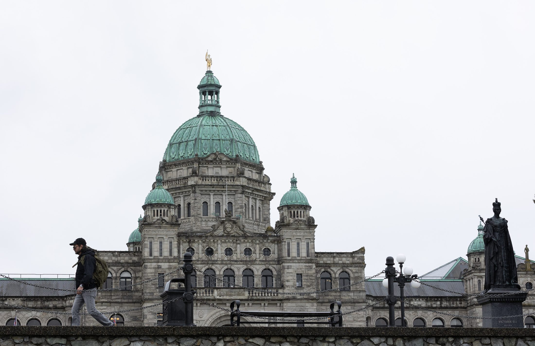 A photo of the BC Legislature taken from the Inner Harbour walkway. A person is walking along the sidewalk above the photographer and the building is silhouetted against a grey sky