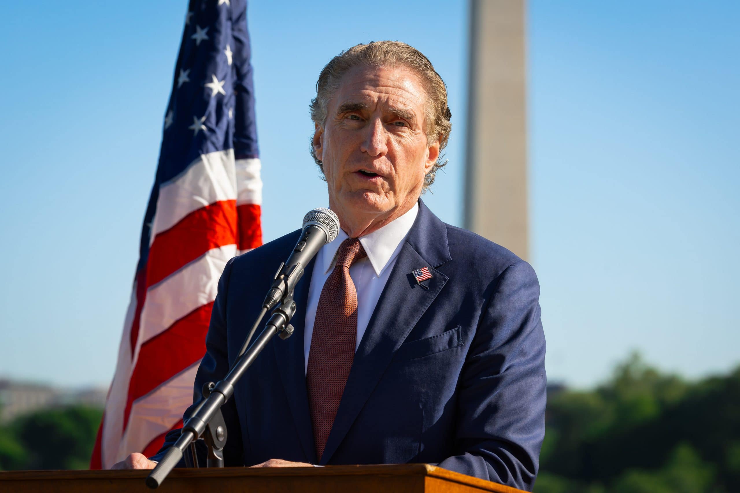 Photo of a man in a blue suit and red tie speaking in front of an American flag
