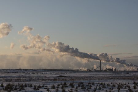 A snowy field with an industrial oil and gas plant in the distance, with smoke billowing into the air.