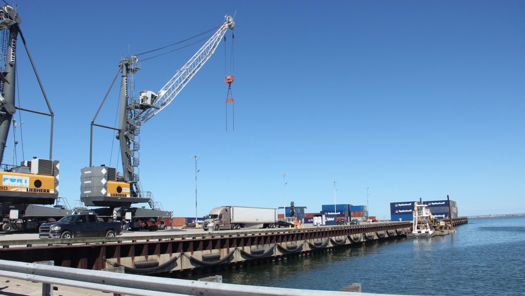 Trucks and cranes are on a wharf jutting out into Lake Erie under a clear blue sky.