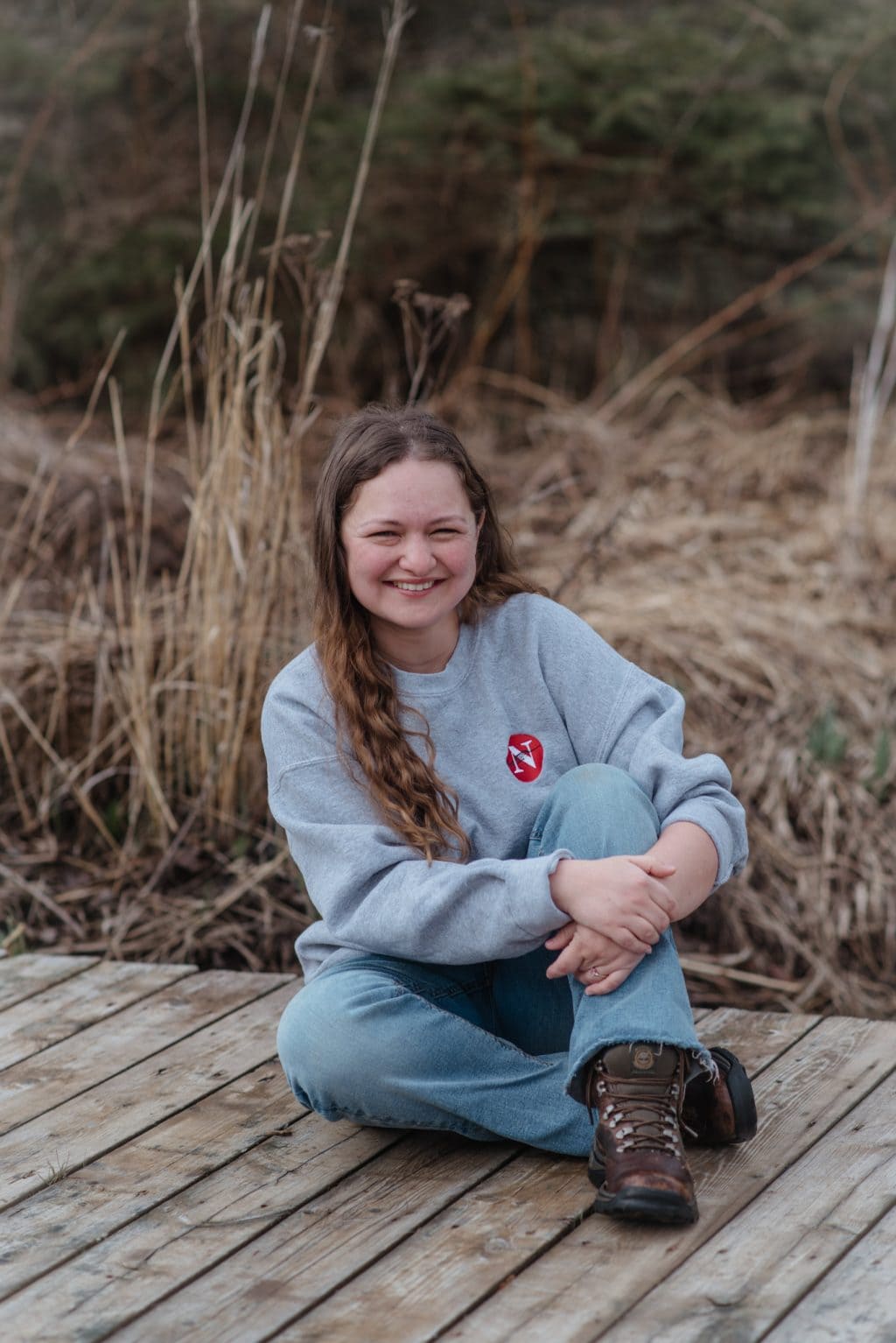 Reporter Leah Borts-Kuperman sits on a small wooden bridge crossing a stream and smiles. She is wearing a sweatshirt with The Narwhal's logo on it.