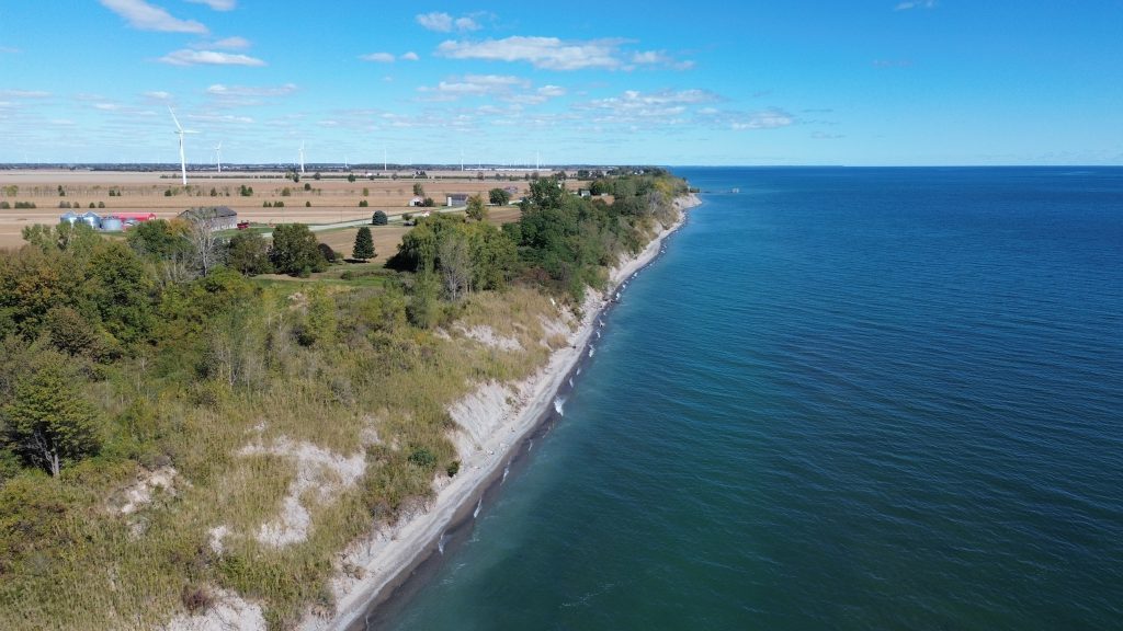A drone photograph of the shore of Lake Erie, with wind turbines on land in the horizon.
