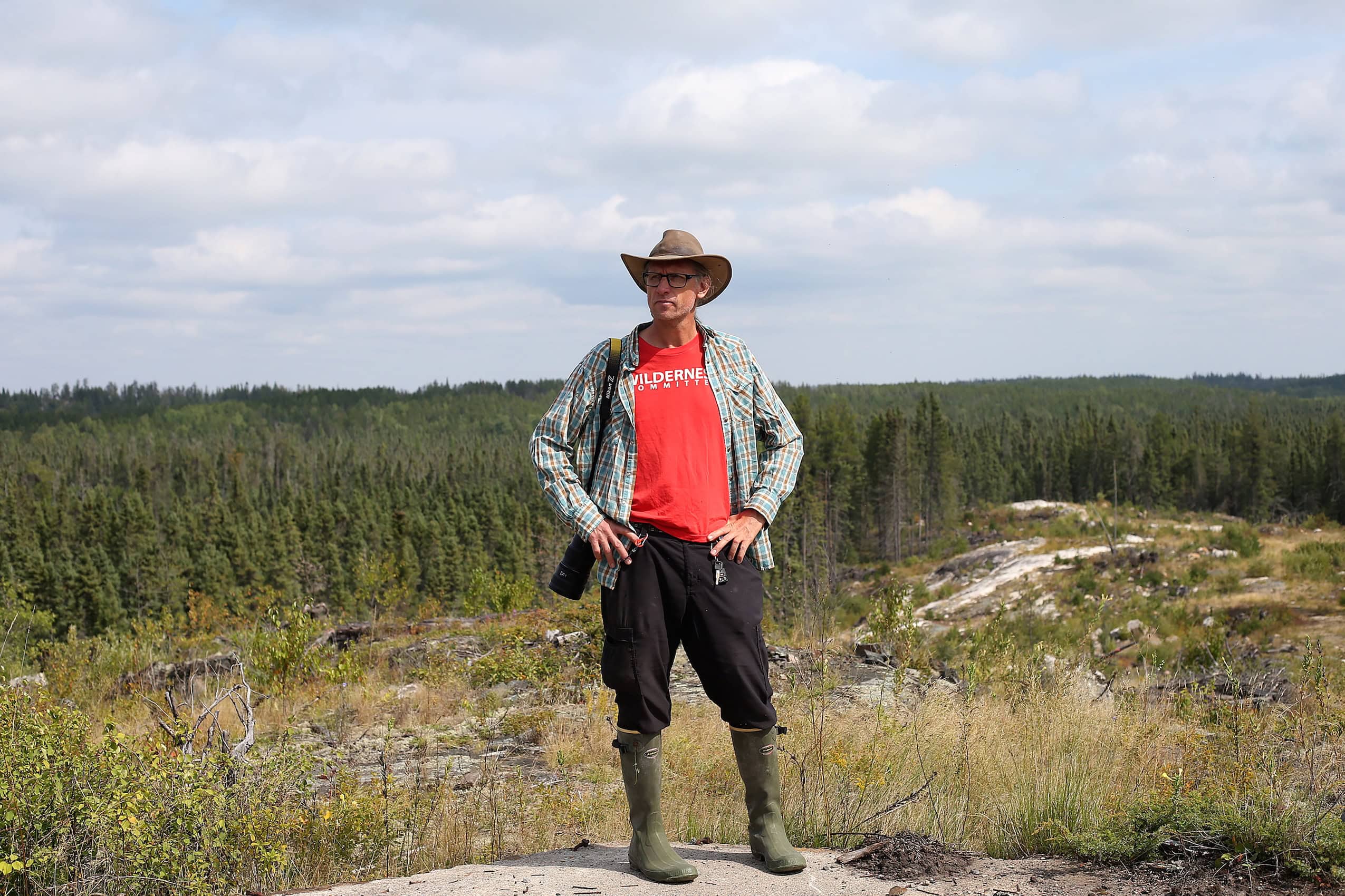 A man wearing a Wilderness Committee t-shirt standing on a rocky outcliff overlooking a forest.