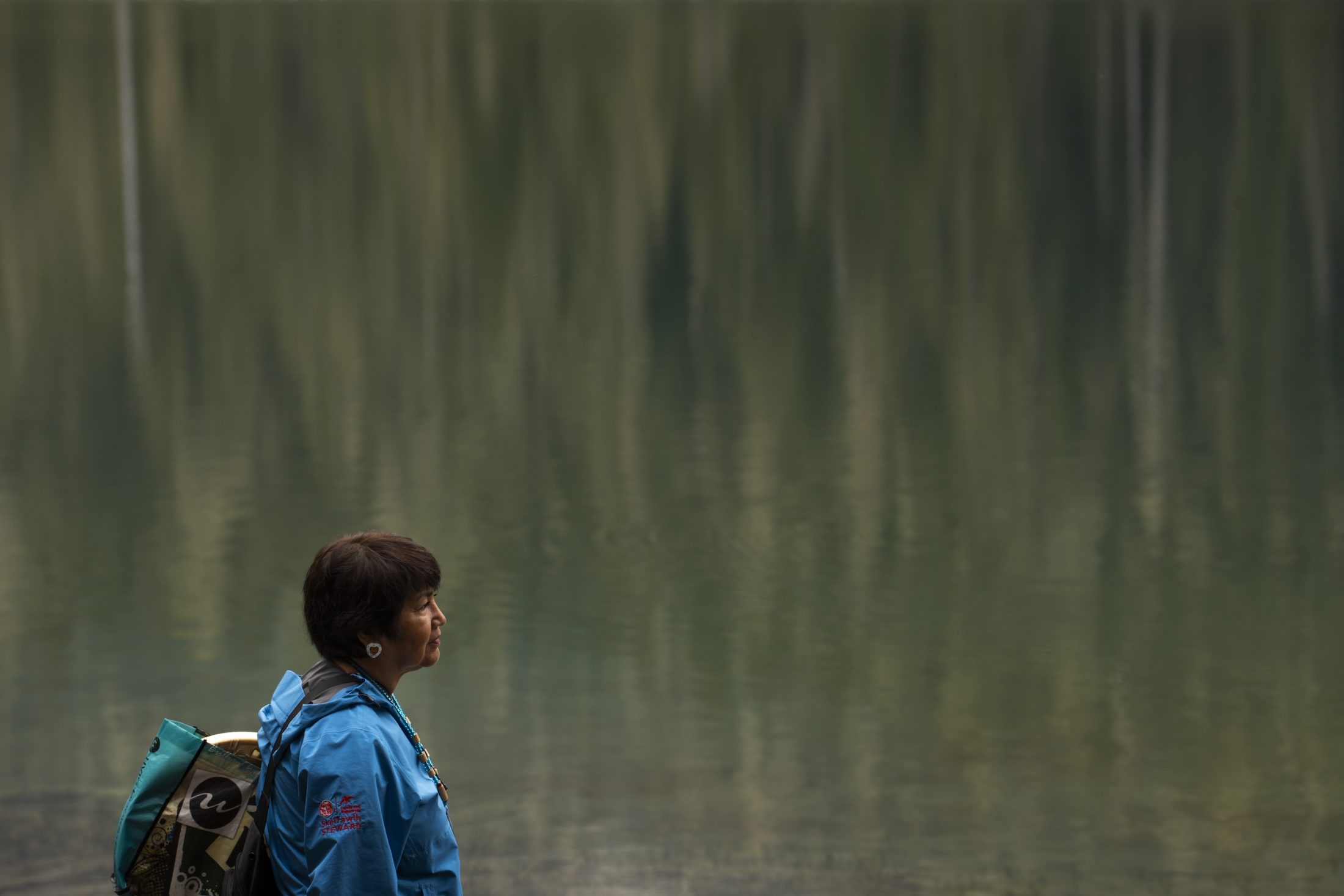 A person stands at the edge of a still lake that is reflecting images of trees.