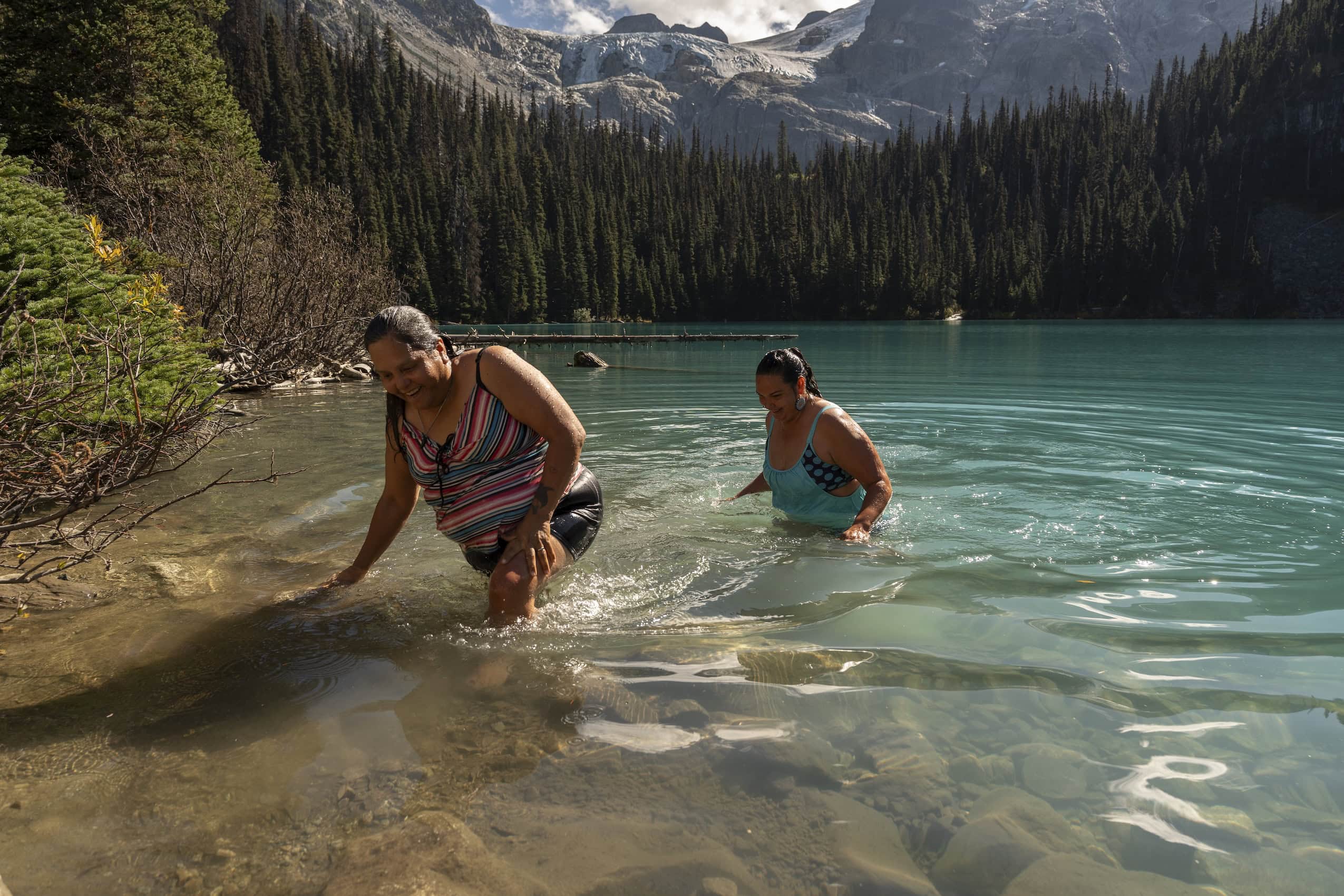 Two women in swimsuits emerge from an emerald green lake, with mountains rising above in the background.