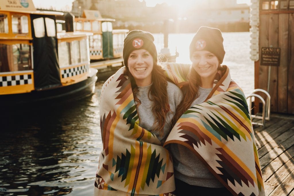 Two women standing on a dock with a little water taxi behind them. They are wrapped in a blanket.