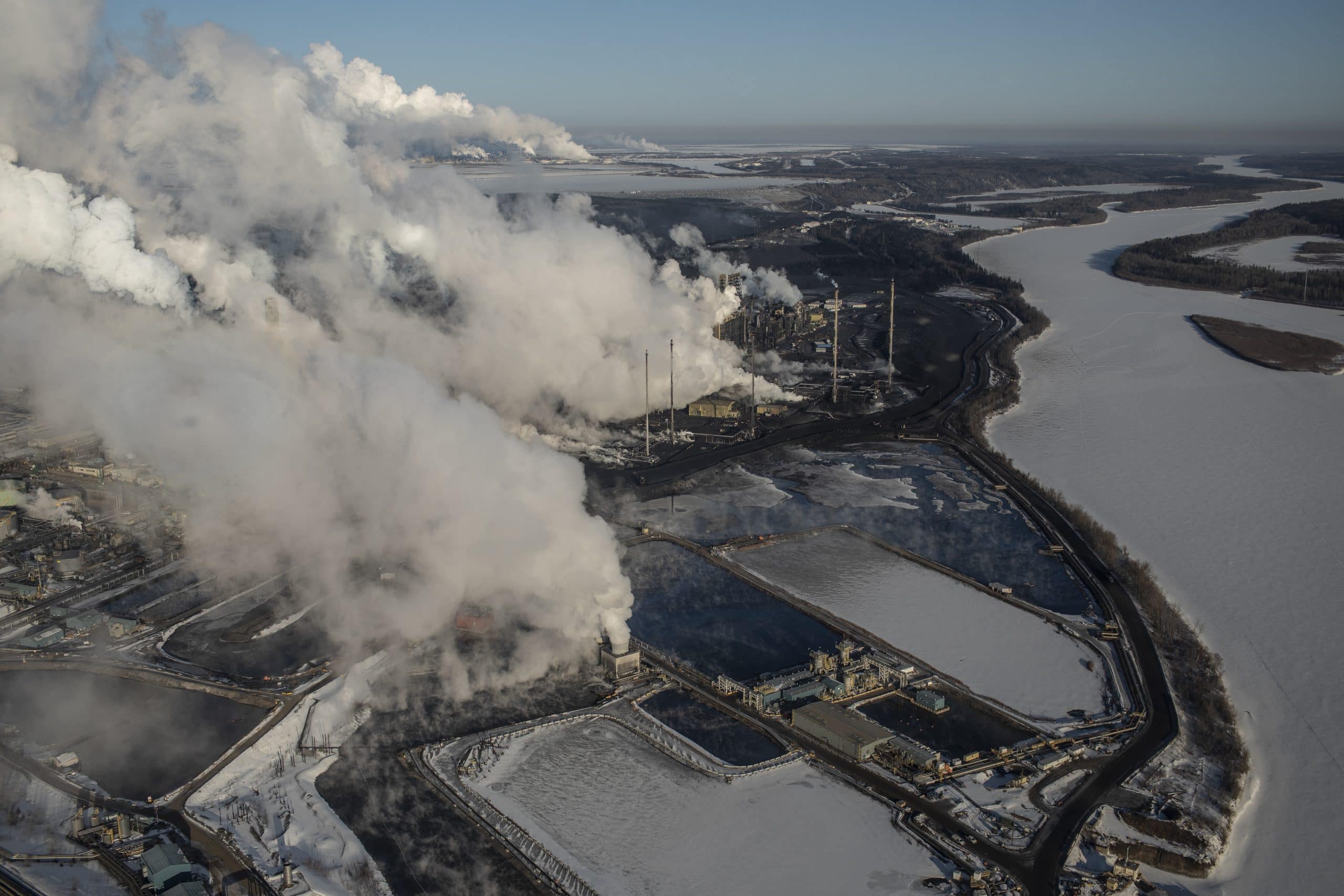 An aerial view of a large oilsands plant with tailings ponds and smoke streaming out of the smokestacks.