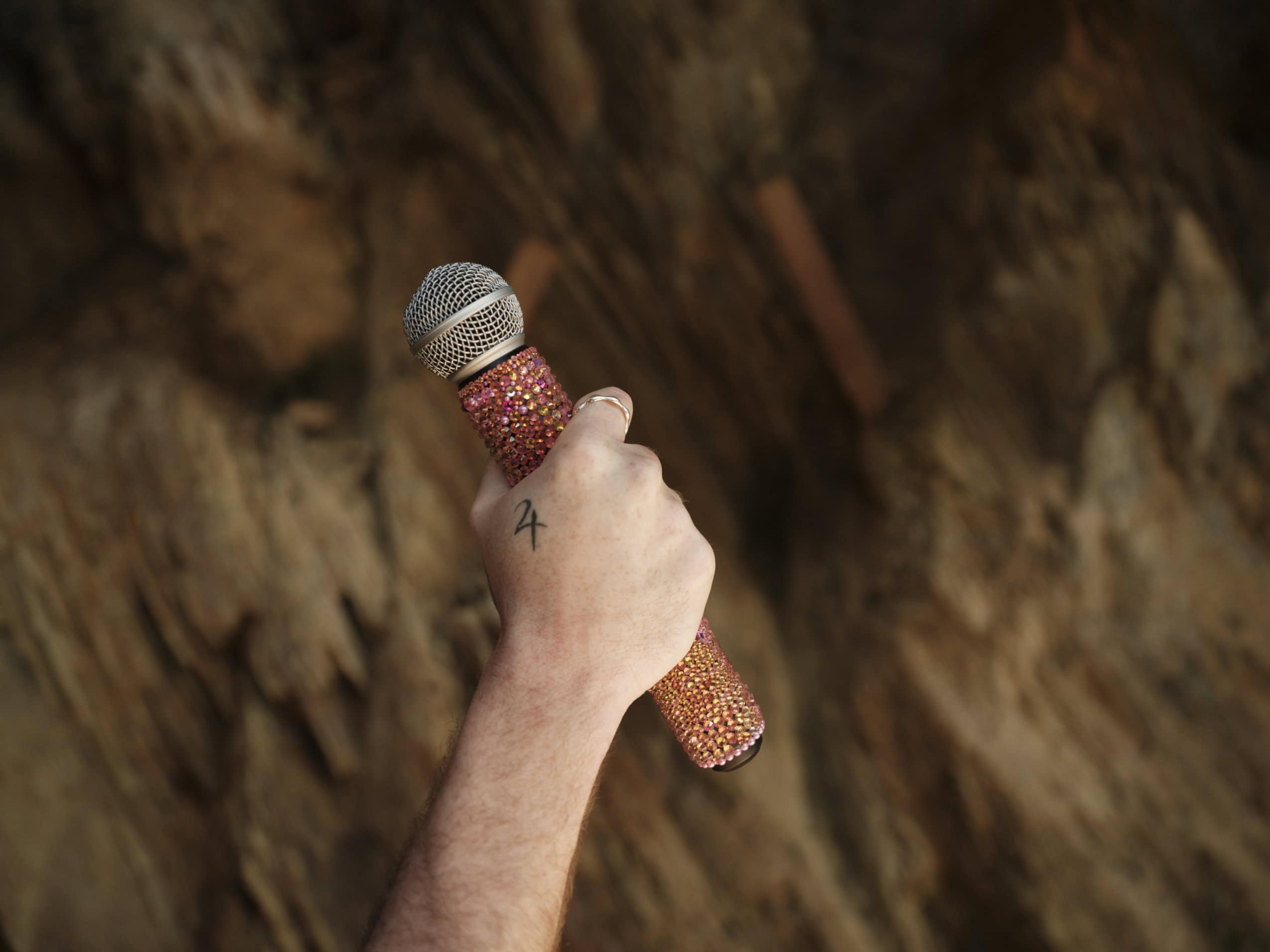 A hand holds a custom jewelled mic in front of a rock wall background.
