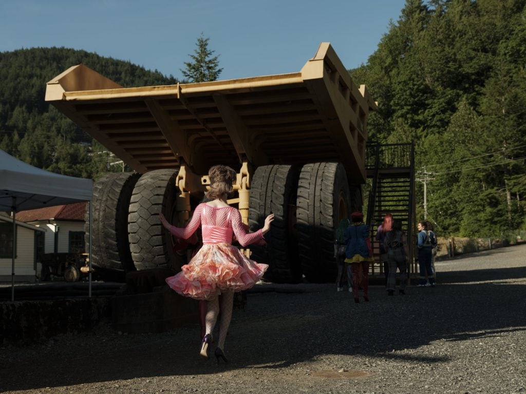 A drag performer in a pink outfit walks toward a large industrial truck.
