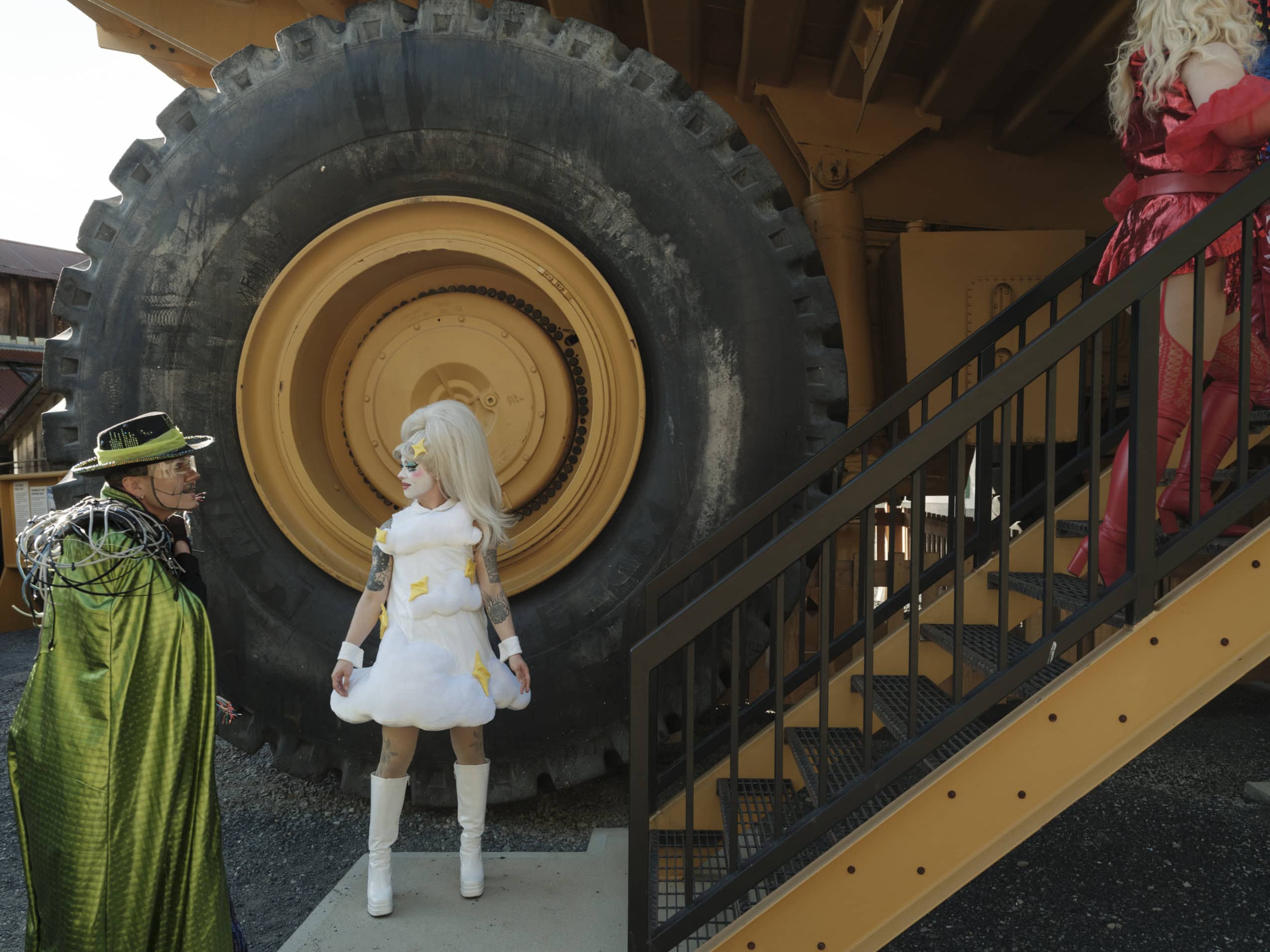 Drag performers in front of a massive industrial truck — the truck's back wheel alone is more than twice the size of a performer standing in front of it.