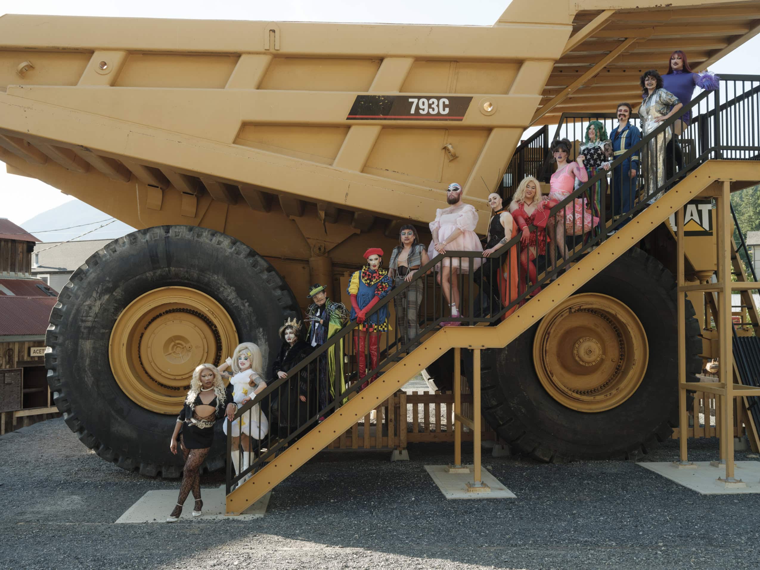 A group of drag artists pose for a photo on a staircase in front of a massive industrial truck.