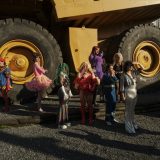 A group of drag artists are dwarfed by a huge industrial truck behind them.