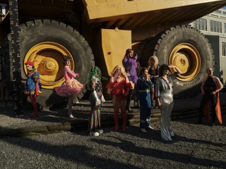 A group of drag artists are dwarfed by a huge industrial truck behind them.