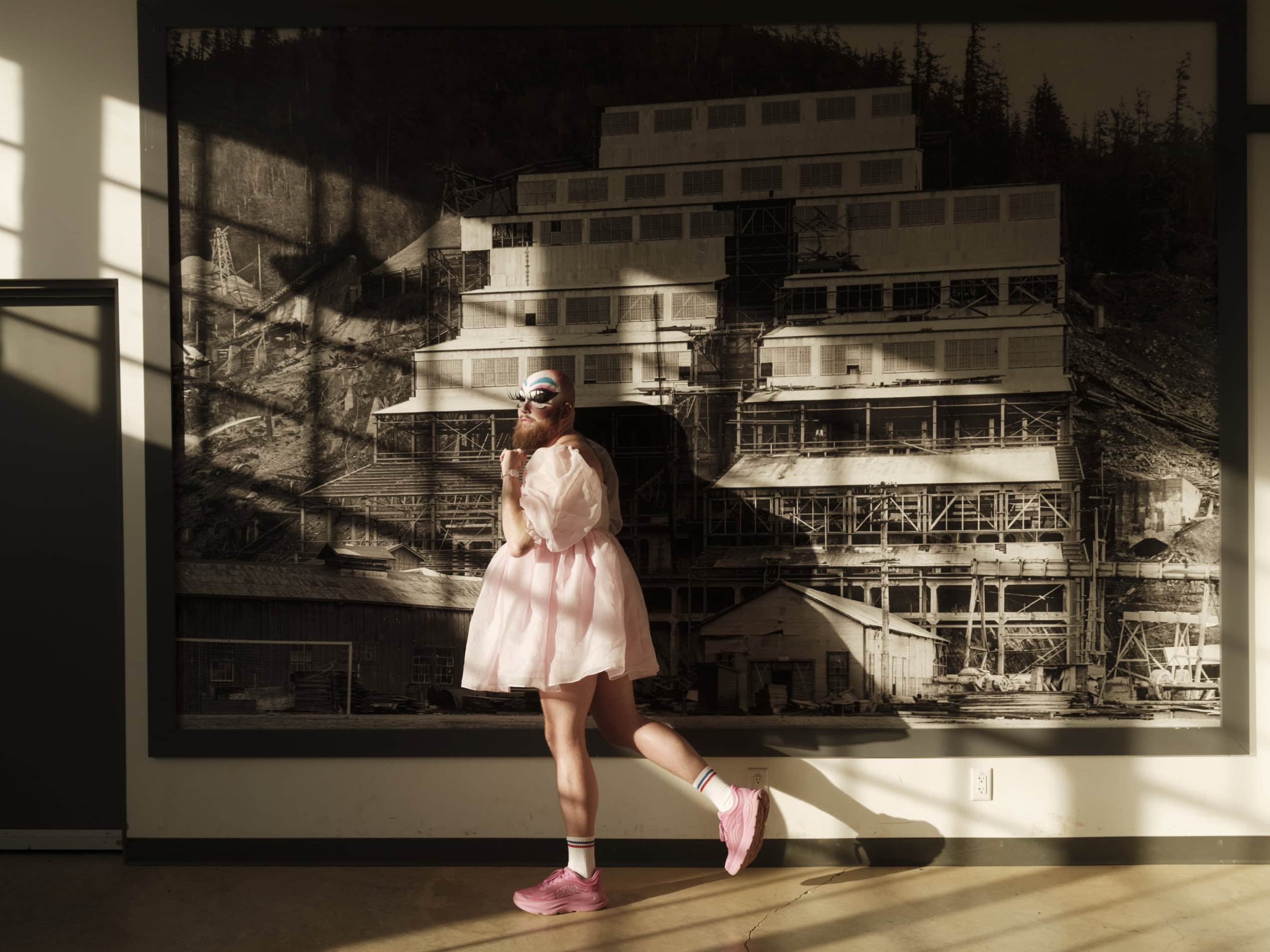 Drag performer Dust Cwaine, wearing a pink outfit and face paint, poses in front of an archival photo of the Britannia Beach copper mine.