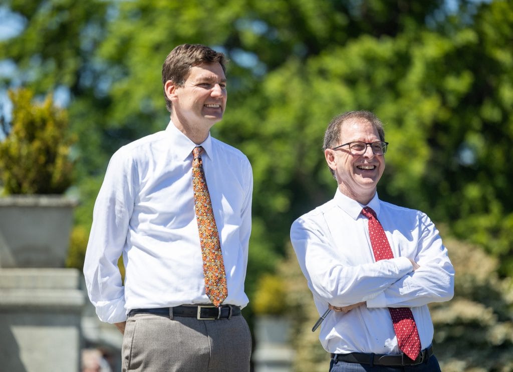B.C. Premier David Eby and Minister of Energy and Climate Solutions Adrian Dix stand in bright sunlight, wearing white shirts and ties.