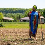 A woman in bright coloured clothing holds a shovel, standing on a farm