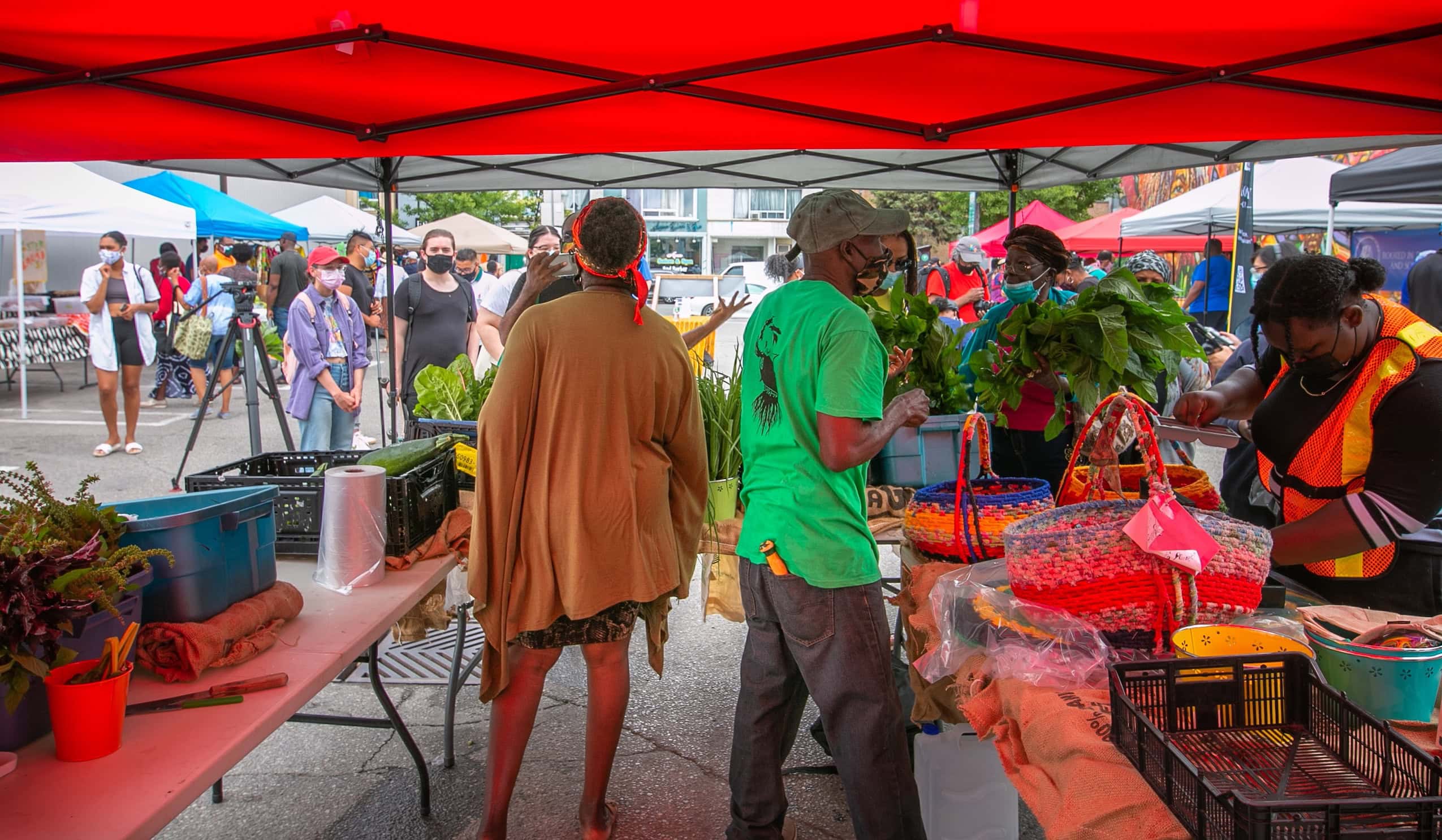 Three Black farmers selling food under a red farmers' market tent.