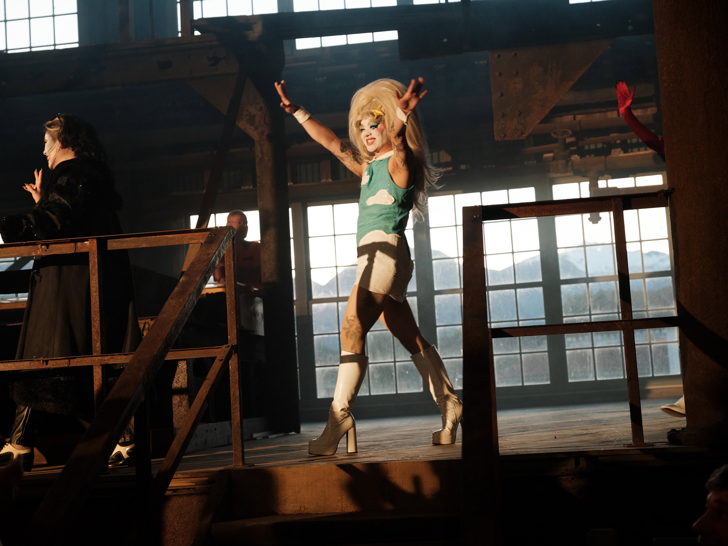 A drag performer raises their hands wearing a blue sequinned dress with clouds on it.