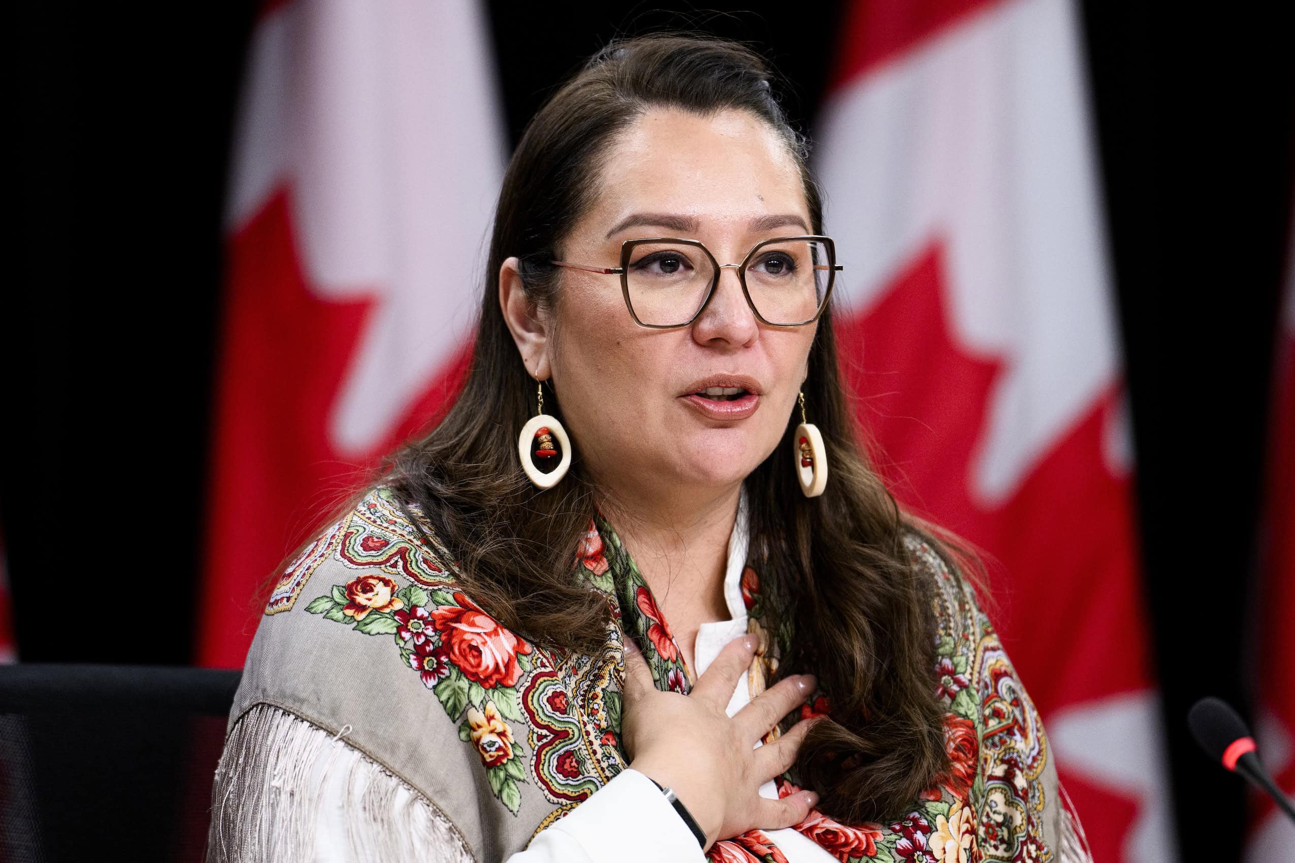Minister of Indigenous Services Mandy Gull-Masty speaks during a press conference. She is wearing glasses, a floral scarf, and statement earrings.