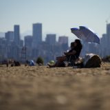 People sit under umbrellas at Locarno Beach in Vancouver. Skyscrapers stand tall in the background.