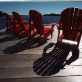 Four red Muskoka chairs sit on a wooden dock overlooking Lake Muskoka on a sunny day.