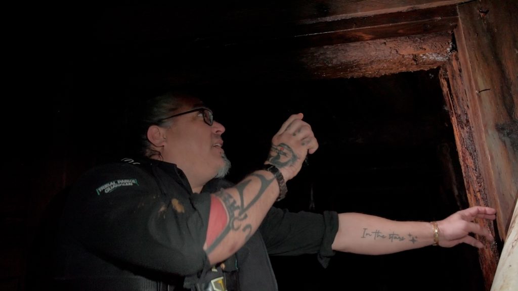A man with tattoos on his arm holds a flashlight to inspect a wooden beam in a dark mine