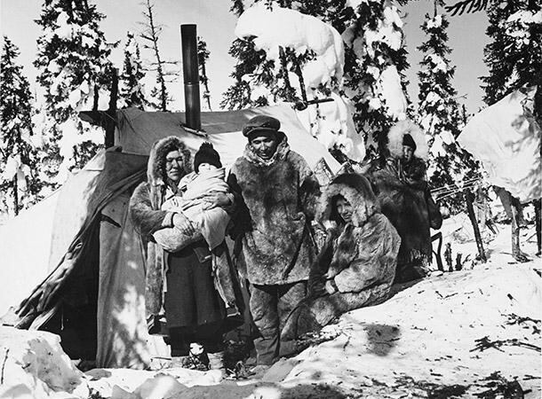 An archival black-and-white image of three Sayisi Dene First Nations people wearing heavy fur coats and hats against a snowy forested background.