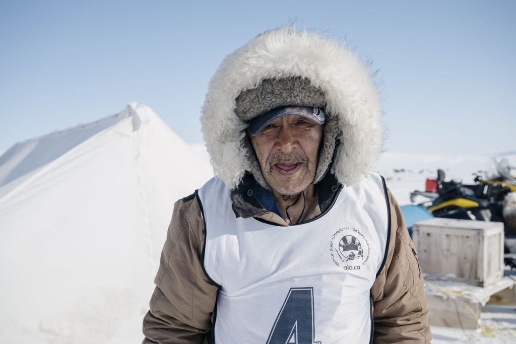 77-year-old Natalino Piugattuk wears winter gear and looks into the camera on a sunny day.