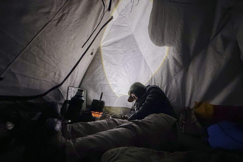A man looks at his phone while boiling water in his tent at night.