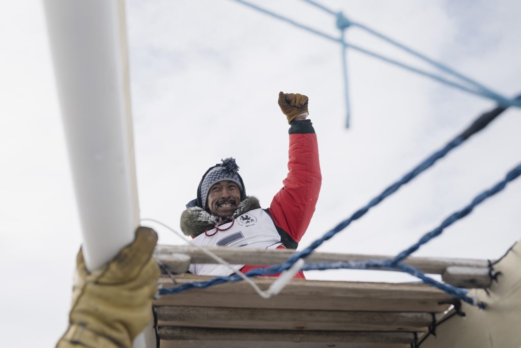 A man wearing winter survival gear hoists his left arm up in celebration, surrounded by a snowy landscape.