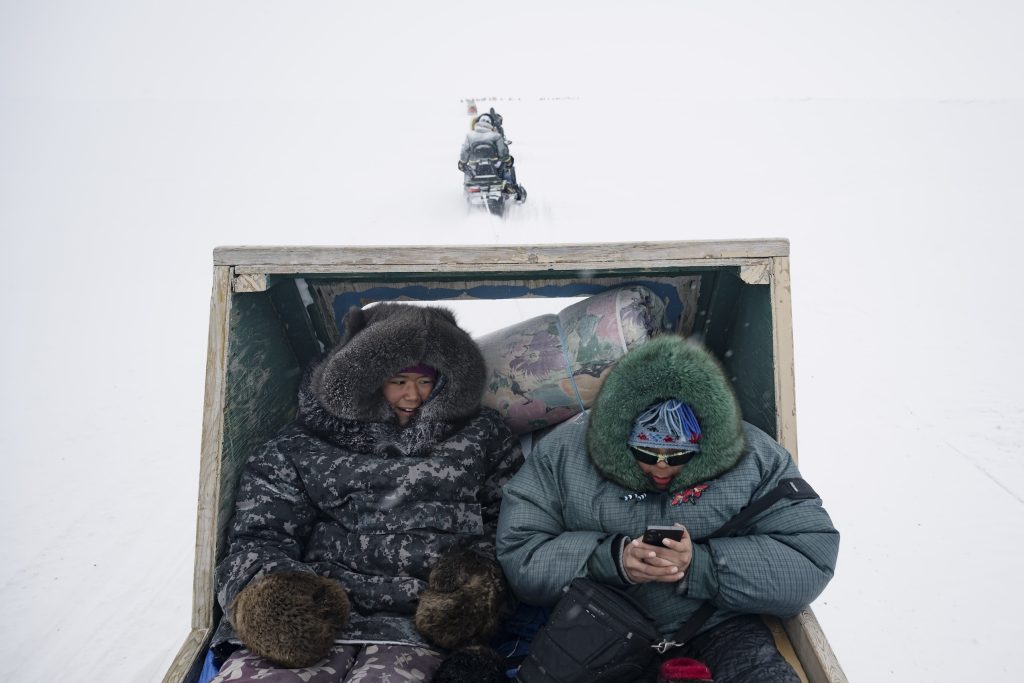 Two young people wearing winter gear look down at a phone while being towed by a snowmobile in a snowy landscape.