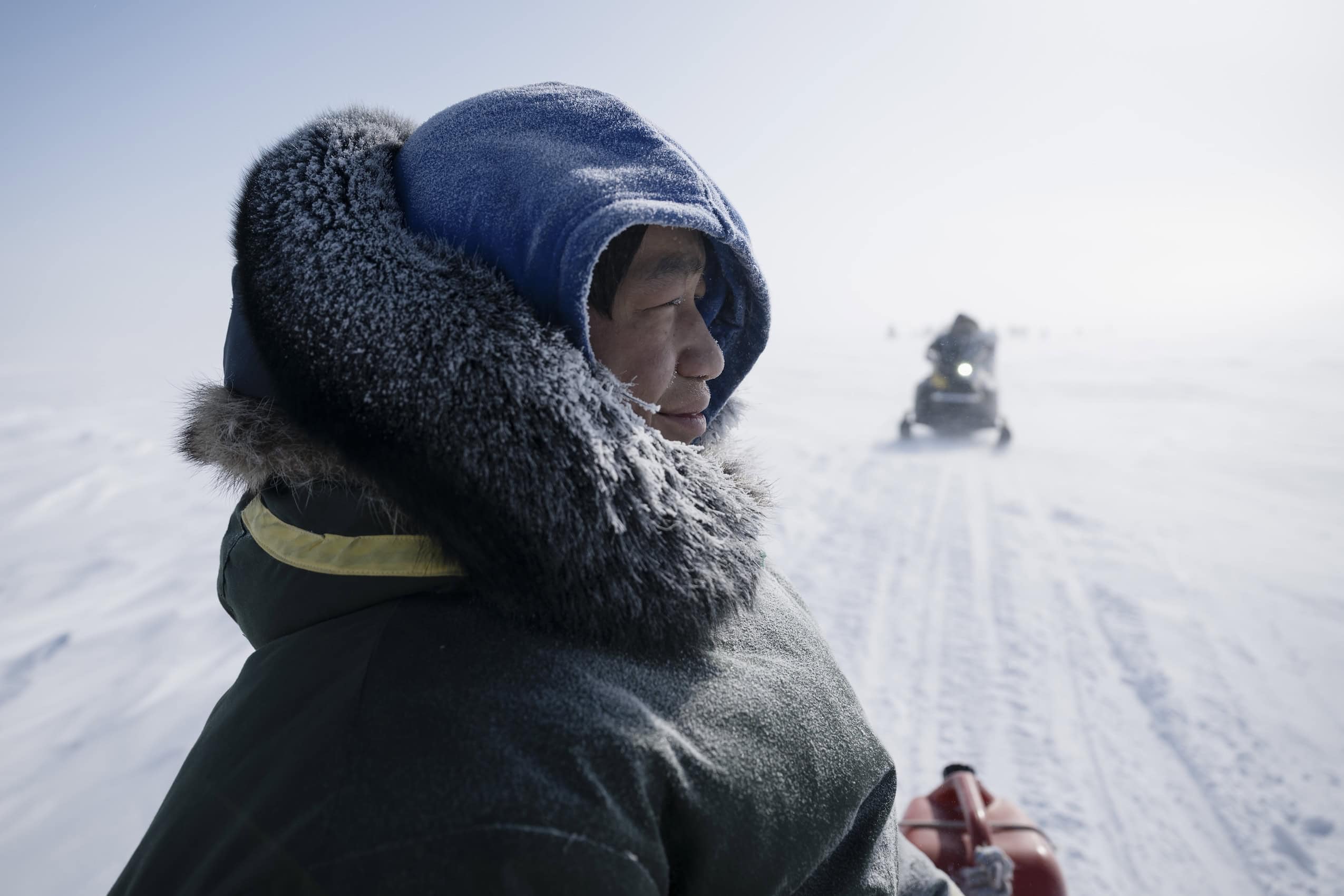 Seen in profile, a person wearing winter sits on a sled being pulled by a snowmobile in the Arctic.