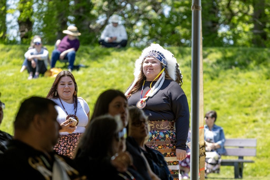 A woman wearing Indigenous regalia stands in the sun and looks at the camera.