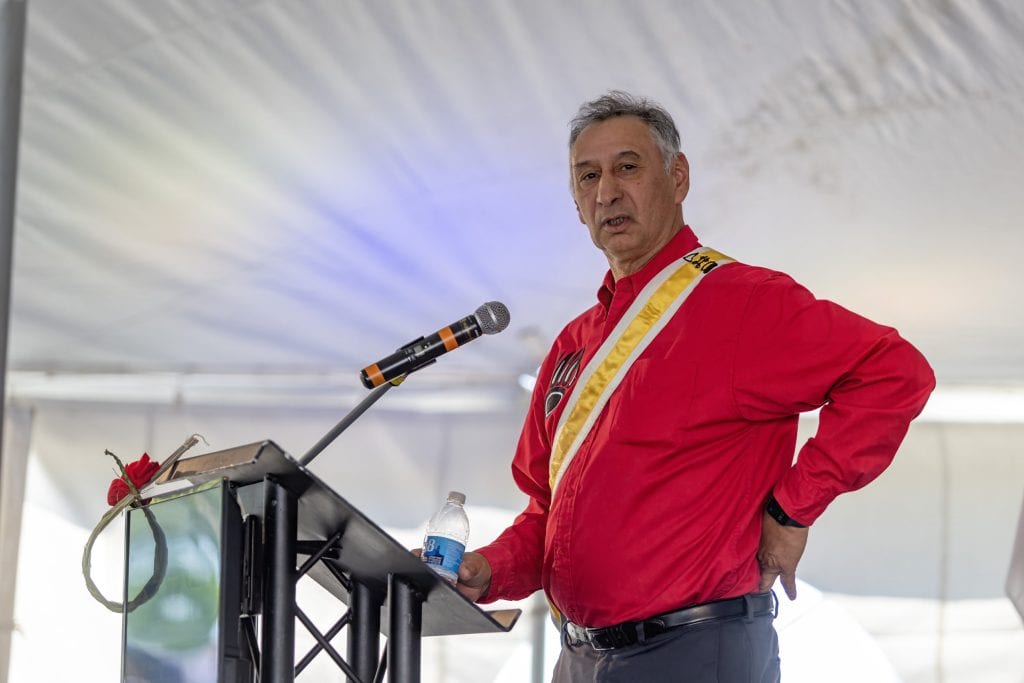 A man wearing a red shirt and a yellow sash speaks into a microphone.