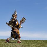 A person wearing Indigenous regalia walks on a grassy field with a blue sky in the background.