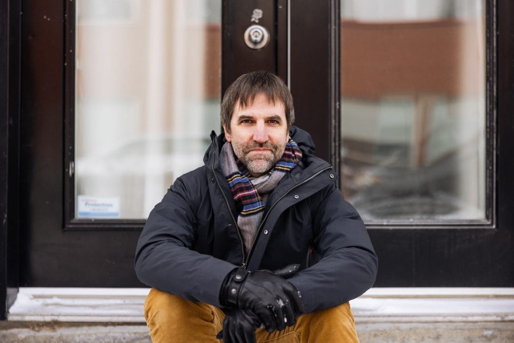 Canadian politician and environmental advocate Steven Guilbeault sits on a doorstep, wearing a scarf and winter coat.