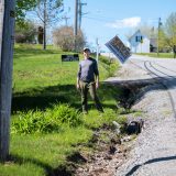 A man stands at the edge of a road, with signs reading "Save Lorneville" standing behind him.