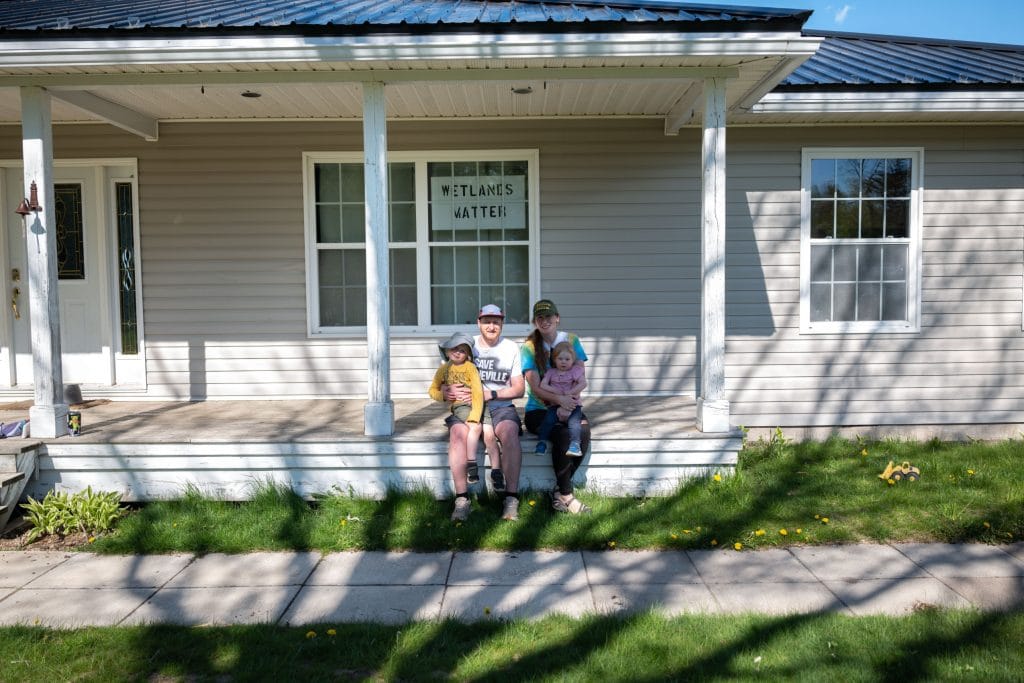 A family sits on their front porch. Behind them, a sign that reads "Wetlands Matter" hangs in a window.