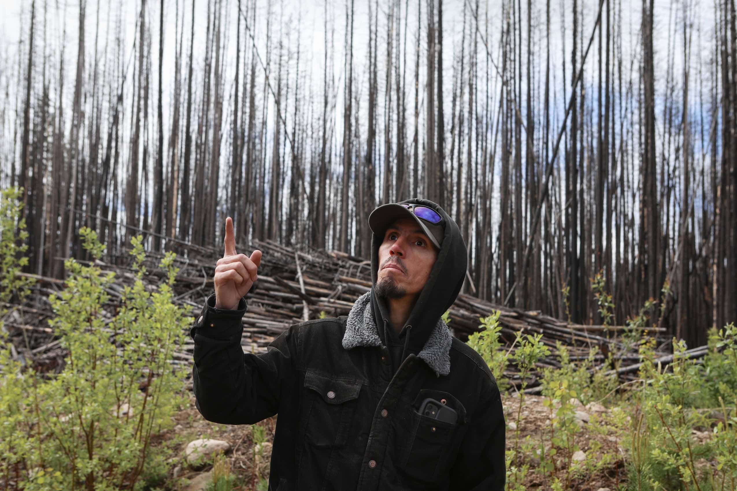 A man wearing a hooded sweatshirt with a wildfire-burned forest behind him.