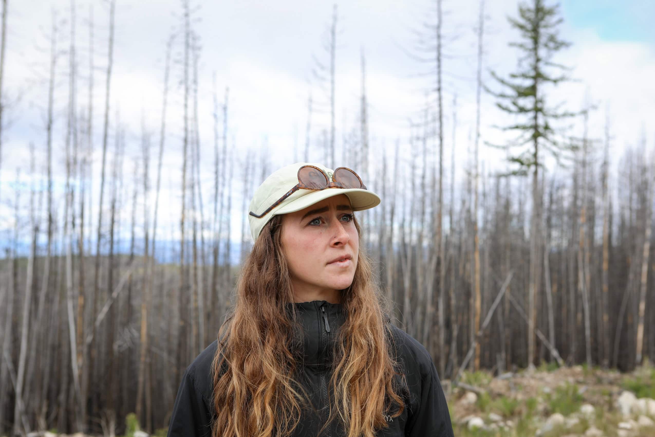 A woman wearing a baseball hat gazes into the distance, with a wildfire-burned forest behind her.