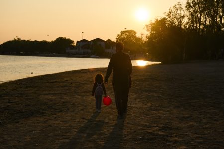 A big, setting sun in a hazy sky, as an adult and child walk along a sandy beach