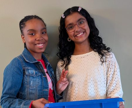 Two girls smile at the camera, one is holding a recycling bin while the other holds up a peace sign