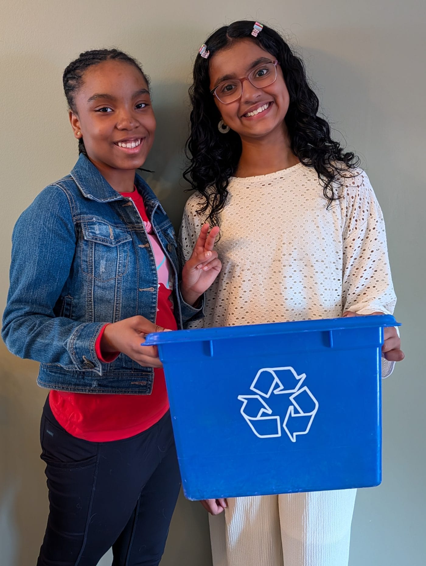 Two girls smile at the camera, one is holding a recycling bin while the other holds up a peace sign