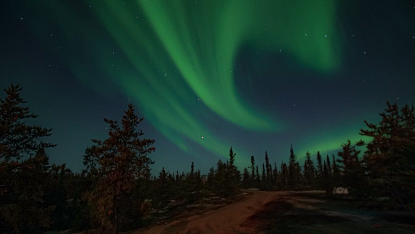 The Northern Lights swirl green in a dark sky over a forested area with a hut in the background.